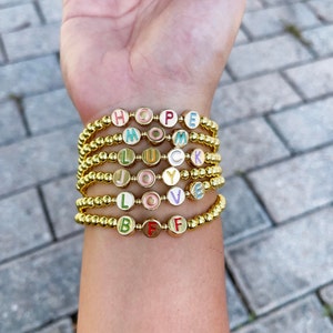 Puede incluir: Una pila de pulseras de cuentas doradas con abalorios de letras de colores que deletrean las palabras "HOPE", "MOM", "LUCK", "JOY", "LOVE" y "BFF".
