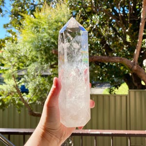 以下が含まれることがあります： 虹色の虹彩を持つ、大きな透明な水晶ポイント。水晶は、緑の葉のぼやけた背景に対して、手で持たれています。