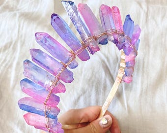 May include: A handcrafted crystal crown featuring a series of colorful quartz crystals in shades of pink, purple, and blue. The crystals are secured with copper wire to a gold-toned headband. The word "Chaeri" is visible in the bottom right corner.