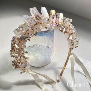 May include: A gold headband with a delicate floral design featuring iridescent crystals, white pearls, and clear rhinestones. The headband is adorned with white and pink crystal-like pieces.