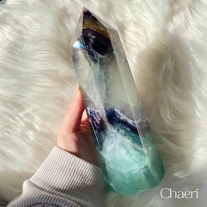 May include: A large, clear fluorite crystal point with green, blue, and purple banding. The crystal is pointed at the top and has a flat base. It is being held in a hand.