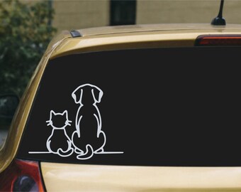 May include: A white vinyl decal sticker featuring the simple line art silhouettes of a cat and a dog sitting side by side, facing away from the viewer. The design is displayed on the rear window of a gold-colored vehicle.