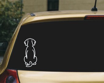 May include: A white vinyl decal featuring the simple line art outline of a dog sitting down, viewed from behind. The sticker is applied to the dark tinted rear window of a gold-colored vehicle, showing the dog's ears, back, and tail.