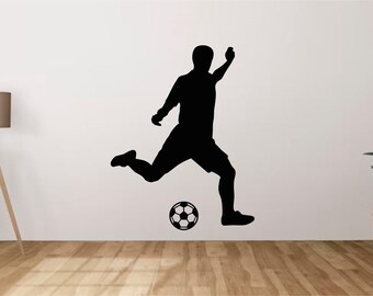 May include: A black wall decal featuring the silhouette of a soccer player in motion, mid-kick, positioned above a detailed black and white soccer ball graphic. The design is rendered in a solid black color, creating a high-contrast visual element.