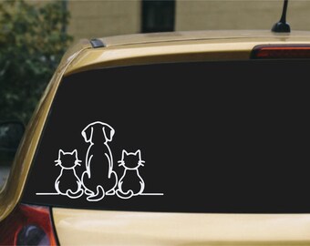 May include: A white vinyl decal sticker applied to a car window, featuring a minimalist line art illustration of a dog sitting in the center flanked by two cats, one on each side, all facing away from the viewer on a single horizontal line.