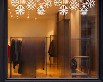 May include: White Christmas ornament decals with snowflake designs are displayed on a storefront window. The ornaments are hanging from white ribbons. The window displays clothing racks and a black door frame.