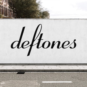 Op de afbeelding: Een witte muur met het woord deftones in een zwart, cursief, gestileerd lettertype. De muur bevindt zich boven een grijze betonnen stoeprand en een donkere asfaltweg met een witte geschilderde lijn, wat een duidelijk contrast oplevert.