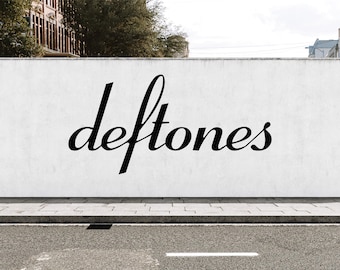 May include: A white wall featuring the word deftones in a black, cursive, stylized font. The wall is positioned above a gray concrete curb and a dark asphalt road with a white painted line, creating a clear contrast between the text and background.