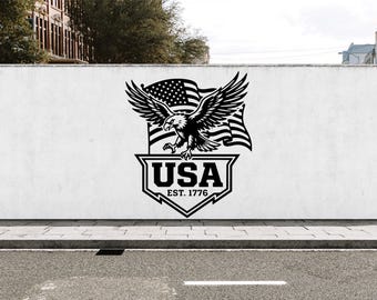 May include: A black graphic decal on a white wall featuring a bald eagle with wings spread wide in front of a waving American flag. Below the eagle is a shield shape containing the bold text USA and the smaller text EST. 1776 centered underneath.
