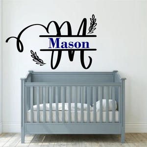 Puede incluir: Una calcomanía de pared en blanco y negro con la letra M y el nombre Mason en un estilo de monograma dividido. La calcomanía está sobre una cuna gris en una guardería.