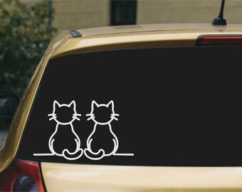 May include: A white vinyl decal featuring the simple line art outlines of two cats sitting side by side, applied to the rear window of a gold-colored car. The design shows the cats from behind with their tails curled and whiskers visible.