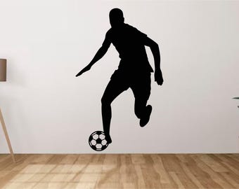 May include: A black wall decal silhouette of a soccer player in motion, mid-stride, with one foot positioned over a classic black and white hexagonal patterned soccer ball. The design is a solid, flat graphic applied to a plain white wall surface.
