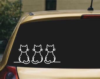 May include: A white vinyl decal featuring a line art illustration of three cats sitting side by side, viewed from behind. The cats are depicted with simple outlines, pointed ears, and curled tails, positioned on a thin horizontal line.