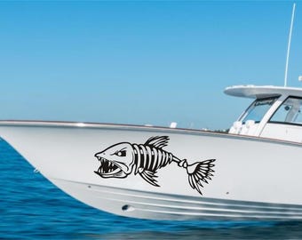 May include: A white boat with a black fish skeleton decal on the side. The fish has a large mouth and sharp teeth. The boat is in the water and the sky is blue.