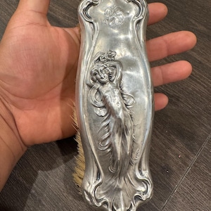 Puede incluir: Un cepillo para el cabello plateado de estilo Art Nouveau con un relieve de una mujer con túnicas fluidas. El cepillo tiene una forma curva y alargada con elementos decorativos florales y fluidos. Las cerdas son de color marrón claro.