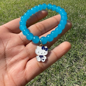 Puede incluir: Una pulsera de cuentas azules con un dije de Hello Kitty. El dije es blanco con un lazo y detalles azules. La pulsera está hecha de cuentas redondas azules translúcidas. La pulsera se muestra en una mano sobre un fondo de hierba verde.
