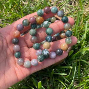 May include: A beaded bracelet featuring a variety of earth-toned gemstones, including green, brown, and tan. The round beads are strung together, creating a natural and organic aesthetic. The bracelet is displayed on a hand against a backdrop of green grass.