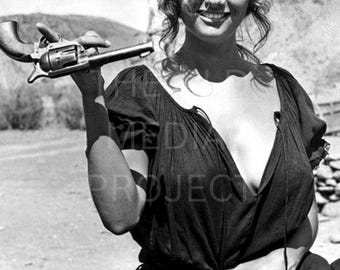 May include: Black and white photograph of a person holding a revolver. The person is wearing a dark dress and a belt with bullets. The background shows a desert landscape.