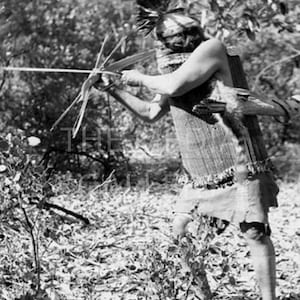 May include: A black and white photograph of a Native American man wearing a feathered headdress and a woven garment, holding a bow and arrow in a forest setting.
