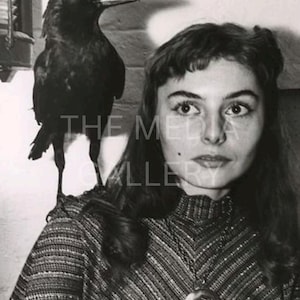 May include: A black and white photo of a woman with a black crow perched on her shoulder. She is wearing a patterned sweater and a necklace with a round pendant.