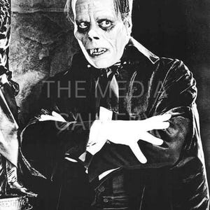 May include: A black and white photo of a man wearing a black suit and a white mask. He has a serious expression on his face and his hands are crossed in front of him.