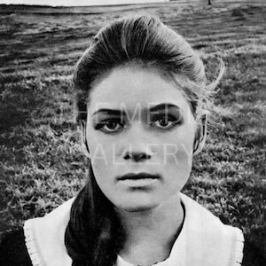 May include: A black and white portrait of a young woman with long, dark hair. She is wearing a white collared shirt with a black trim. The background is a field of grass.