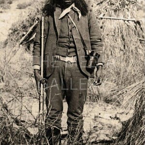 May include: A black and white photograph of a man wearing a hat and holding a rifle. He is standing in a field of tall grass.