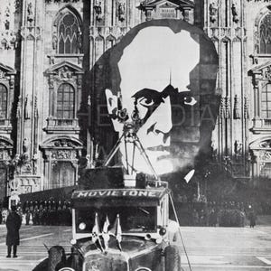 May include: A black and white photo of a large banner with a man's face on it. The banner is hanging from a building. A black car with the word "MOVIETONE" on the side is in the foreground. The car is decorated with flags.