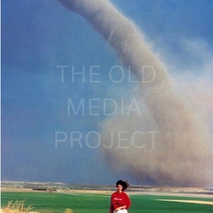 May include: A person stands on a road, observing a large tornado in a rural landscape. The sky is a mix of blue and gray, with the tornado dominating the scene. The text "THE OLD MEDIA PROJECT" is overlaid on the image.
