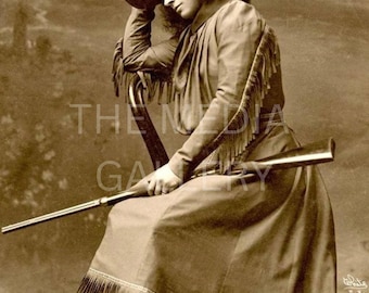 May include: A woman in a cowboy hat and fringed outfit sits on a chair holding a rifle. The photo is in sepia tone.