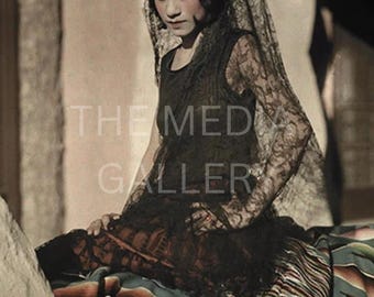 May include: A vintage-style photograph of a person wearing a black lace dress and veil, adorned with a red flower. The person is seated on a colorful striped blanket. The image has a sepia tone, evoking a sense of nostalgia.