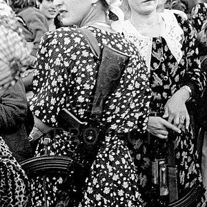 May include: Black and white image of several people, some wearing floral print dresses and white headscarves. Each person is carrying a rifle. The image is a medium shot, with the subjects in the foreground.