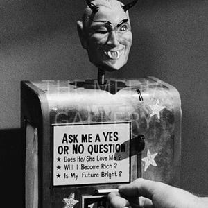 May include: A black and white photo of a fortune teller machine with a devil head on top. The machine has a sign that reads "Ask me a yes or no question." The sign also lists three questions: "Does He/She Love Me?", "Will I Become Rich?", and "Is My Future Bright?"
