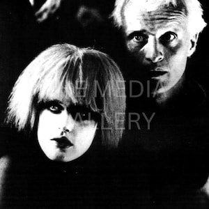 May include: Black and white portrait featuring two individuals. One, with short, light-colored hair, gazes directly at the viewer. The other, with a similar hairstyle, is positioned behind, also looking forward. The image has a dramatic, high-contrast lighting.