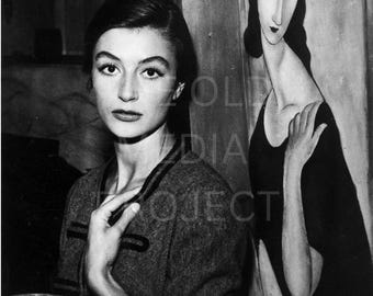 Può includere: Fotografia in bianco e nero di una donna con una giacca scura, in posa con un dipinto di una donna con un vestito nero. La donna nella foto guarda la fotocamera.