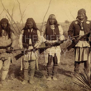 May include: Four Native American men in traditional clothing stand in a field holding rifles. They are wearing feathered headdresses and have long hair. The men are standing in front of a backdrop of trees and bushes.