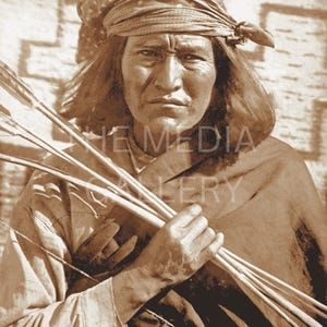 May include: A Native American man wearing a patterned bandana and traditional clothing holds a bundle of arrows in his right hand. He has a serious expression on his face.
