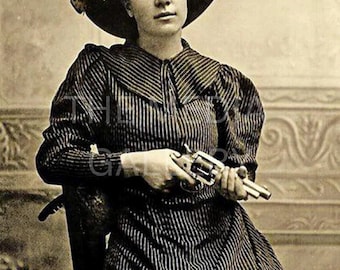 May include: A woman in a striped dress and a wide-brimmed hat holds a revolver in her hands. The photo is in black and white and has a vintage feel.