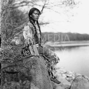 May include: A young person wearing traditional clothing sits on a rock by a body of water. They have long, braided hair and are looking off to the side.