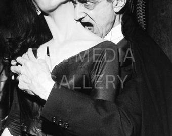 May include: Black and white photograph of a man in a suit biting a woman's neck. The woman is wearing a dark dress and has dark hair and makeup. The man has dark hair and a stern expression. The image has a vintage, gothic theme.