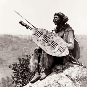May include: A black and white photograph of a man holding a shield and spear. He is wearing a traditional outfit and is sitting on a rock.