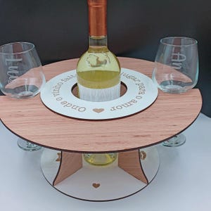 May include: A wooden wine holder with two glass cups and a bottle of white wine. The holder has a white circle in the center with the text "Onde o amor e o vinho faltam, lugar para o amor" and a heart.