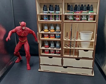 Puede incluir: Un organizador de almacenamiento de madera con múltiples estantes y cajones para guardar botellas de pintura y pinceles. El organizador tiene un asa en la parte superior y está hecho de madera clara. Hay varias filas de pequeñas botellas de pintura en el organizador, así como una taza de plástico blanca.