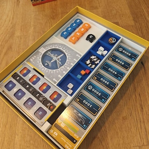 May include: A board game set in a yellow tray, containing various game components. These include blue and orange dice, black dice, and cards with numbers and tokens. The game box, with an airplane graphic, is visible in the background.