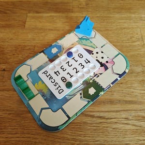 May include: A white game board with a discard pile, a blue house token, and a number tracker with green and blue tokens. The numbers on the tracker go from 0 to 4.