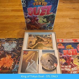 Puede incluir: Juego de mesa "King of Tokyo Duel" con una caja que muestra ilustraciones de monstruos. El juego incluye tableros, cartas y un libro de reglas. Los componentes del juego se muestran sobre una superficie de madera.