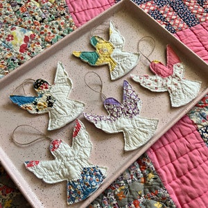 Puede incluir: Cinco adornos de p&aacute;jaros de tela hechos a mano con varias alas estampadas, incluyendo dise&ntilde;os florales y geom&eacute;tricos, descansan sobre una bandeja rosa moteada. Los p&aacute;jaros son blancos con acentos coloridos y tienen lazos de cuerda para colgar.