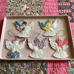 Puede incluir: Seis adornos de &aacute;ngel hechos a mano con cuerpos acolchados blancos y alas y acentos de tela colorida. Cada &aacute;ngel tiene un dise&ntilde;o de ala con diferentes patrones, incluidos patrones florales y geom&eacute;tricos. Se exhiben en una bandeja rosa.