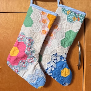 Puede incluir: Dos calcetines navideños acolchados confeccionados con un mosaico de piezas de tela hexagonales. Los calcetines presentan una variedad de telas florales y de colores sólidos en tonos rosa, azul, amarillo, verde y blanco, con un borde a rayas azules y blancas.