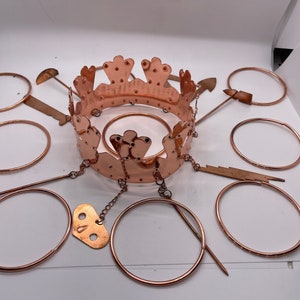 Può includere: Una corona color rame con un design decorativo, circondata da diversi anelli ed elementi decorativi color rame. La corona presenta un motivo dettagliato e ritagliato ed è collegata a un ciondolo a forma di chiave tramite una catena. Gli oggetti sono disposti su una superficie bianca.
